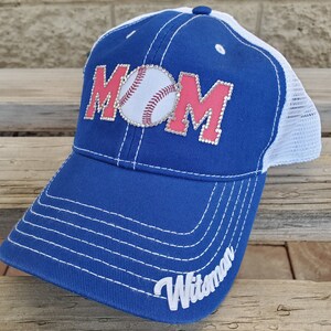 May include: A blue and white baseball cap with a pink "MOM" design featuring a rhinestone baseball. The cap has a white mesh back and the word "Witman" embroidered on the side.
