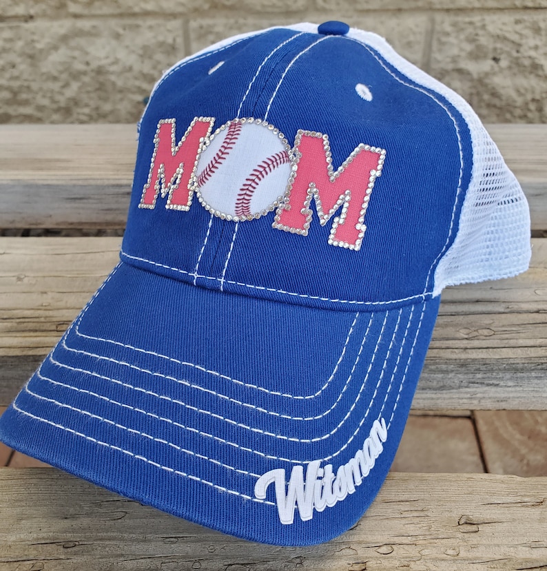 May include: A blue and white baseball cap with a pink "MOM" design featuring a rhinestone baseball. The cap has a white mesh back and the word "Witman" embroidered on the side.