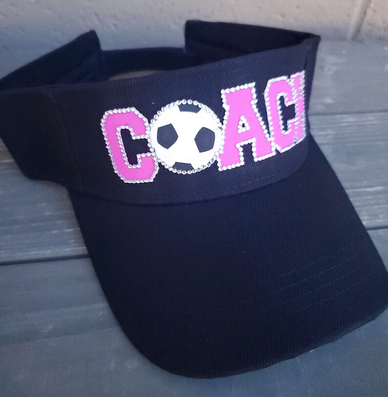 May include: Black visor with a pink "COACH" design and a white soccer ball in the center. The letters are outlined with rhinestones.