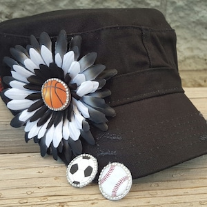 May include: Black canvas baseball cap with a white and black flower embellishment. The flower has a basketball in the center. The cap has a distressed look. There are two earrings with a soccer ball and a baseball design.