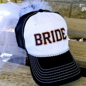 May include: White and black baseball cap with a white tulle veil attached. The cap has a black mesh back and the word "BRIDE" in black with orange trim and rhinestones on the front.