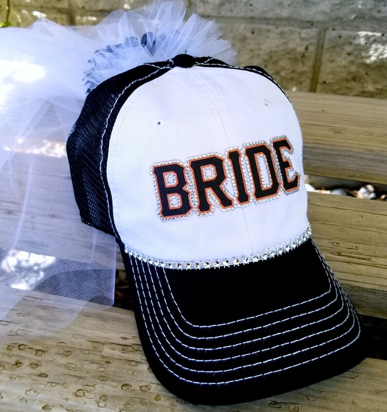 May include: White and black baseball cap with a white tulle veil attached. The cap has a black mesh back and the word "BRIDE" in black with orange trim and rhinestones on the front.