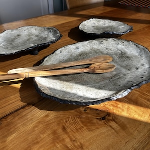 May include: Three irregular, oval-shaped ceramic platters with a mottled grey and white finish and dark, textured edges. A wooden serving spoon and a wooden serving fork are placed diagonally across the largest platter in the foreground.