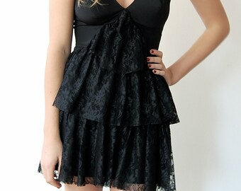 May include: A woman wearing a black layered lace dress with a beaded necklace. The dress has a V-neckline and a ruffled skirt.