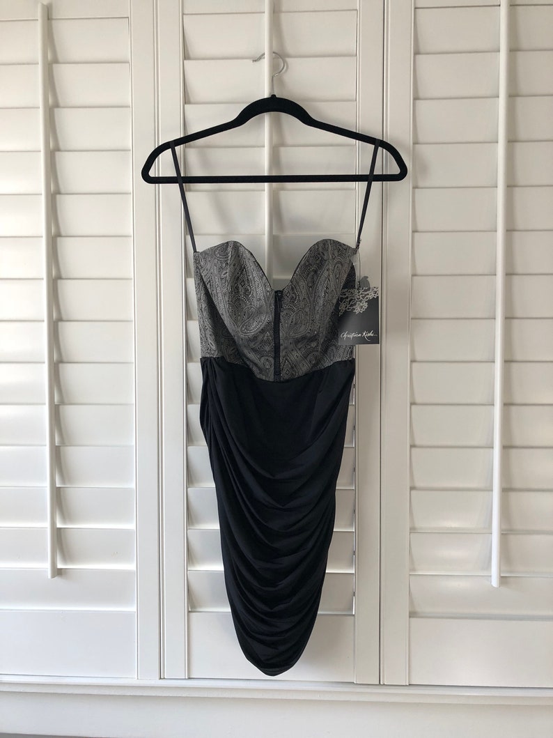 May include: A black and silver strapless dress with a fitted bodice and a ruched skirt. The bodice has a paisley pattern and the skirt is made of a stretchy fabric. The dress has a tag that says "Christian Audigier".