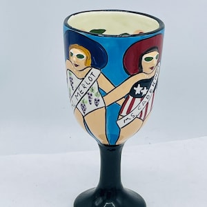 May include: A hand-painted wine glass with a black stem and base. The glass features two figures in swimsuits and hats, one with a "MERLOT" sash and the other with a "MS. CABERNET" sash. The background is blue.