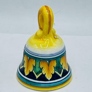May include: A small, yellow ceramic bell with a blue, green, and yellow leaf design. The bell has a loop handle.