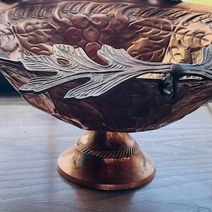 May include: A copper-toned decorative bowl with a pedestal base. The bowl features embossed floral and leaf patterns. The rim has decorative leaf-shaped handles. The bowl is likely made of metal and is suitable for display or serving.