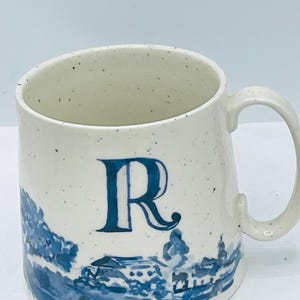 May include: Cream-colored ceramic mug with a blue letter "R" and a blue landscape design. The mug has a speckled finish and a curved handle. The landscape design features buildings and trees.