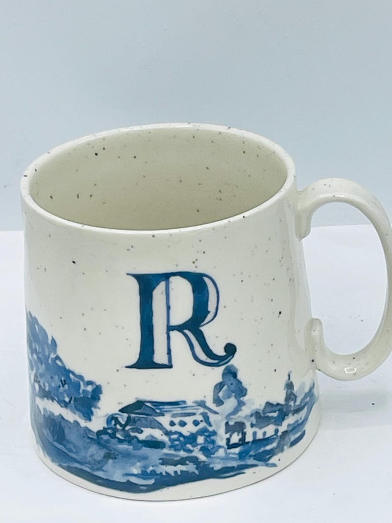 May include: Cream-colored ceramic mug with a blue letter "R" and a blue landscape design. The mug has a speckled finish and a curved handle. The landscape design features buildings and trees.
