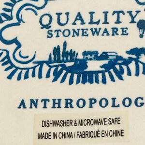 May include: Close-up of a white stoneware product with blue decorative elements. The text "QUALITY STONEWARE" is visible, along with the word "ANTHROPOLOGIE". A sticker indicates the product is dishwasher and microwave safe.