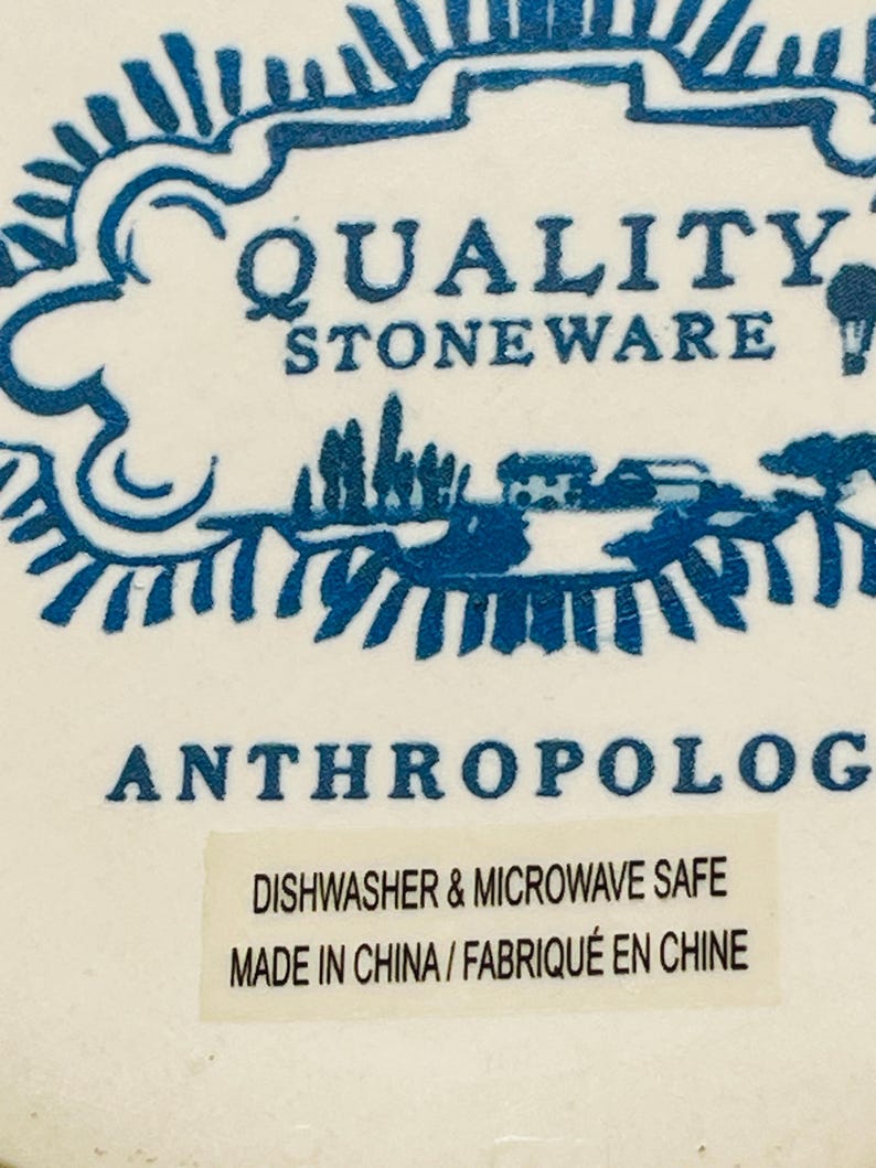 May include: Close-up of a white stoneware product with blue decorative elements. The text "QUALITY STONEWARE" is visible, along with the word "ANTHROPOLOGIE". A sticker indicates the product is dishwasher and microwave safe.