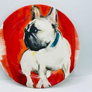 May include: Round decorative plate featuring a watercolor painting of a French Bulldog. The dog is white with black markings and a blue collar, set against a vibrant red background. A unique piece for dog lovers.