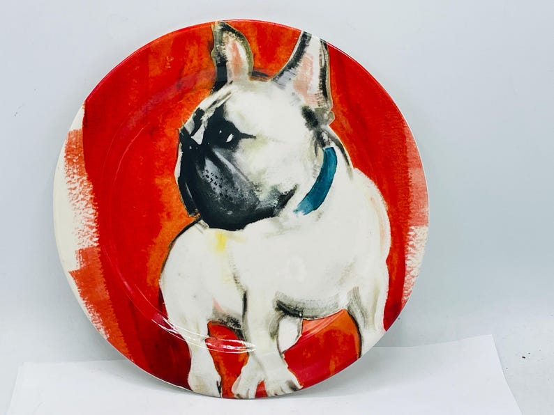 May include: Round decorative plate featuring a watercolor painting of a French Bulldog. The dog is white with black markings and a blue collar, set against a vibrant red background. A unique piece for dog lovers.