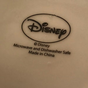 Puede incluir: Taza de cer&aacute;mica blanca con un logotipo de Disney negro y el texto "Microwave and Dishwasher Safe Made in China".