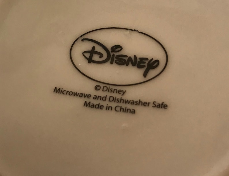 Puede incluir: Taza de cer&aacute;mica blanca con un logotipo de Disney negro y el texto "Microwave and Dishwasher Safe Made in China".