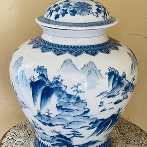 May include: A white and blue ceramic jar with a lid. The jar features a landscape scene in blue, with mountains, trees, and buildings. The lid and base have floral and geometric patterns. The jar is a decorative item.