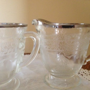 May include: A vintage glass creamer and sugar bowl set with a silver rim and an etched floral design.