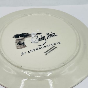 May include: Off-white ceramic plate with a red rim. The underside features the text "Sally Muir for ANTHROPOLOGIE" and other text. The plate is round and has a slightly raised edge.