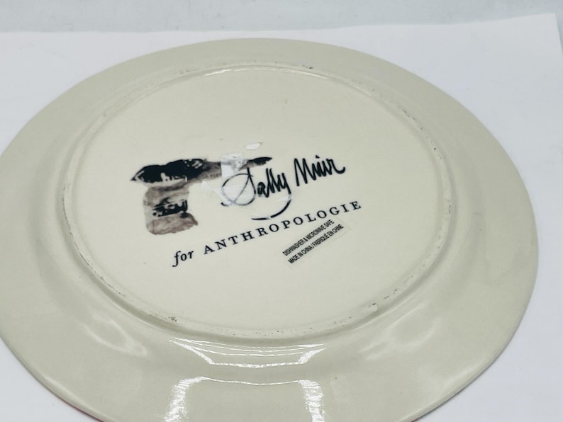 May include: Off-white ceramic plate with a red rim. The underside features the text "Sally Muir for ANTHROPOLOGIE" and other text. The plate is round and has a slightly raised edge.