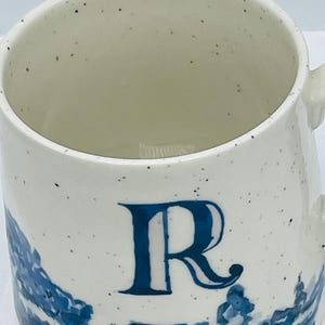 May include: White ceramic mug with a blue letter "R" and a blue landscape design. The mug has a speckled finish and a handle. The design is reminiscent of classic blue and white pottery.