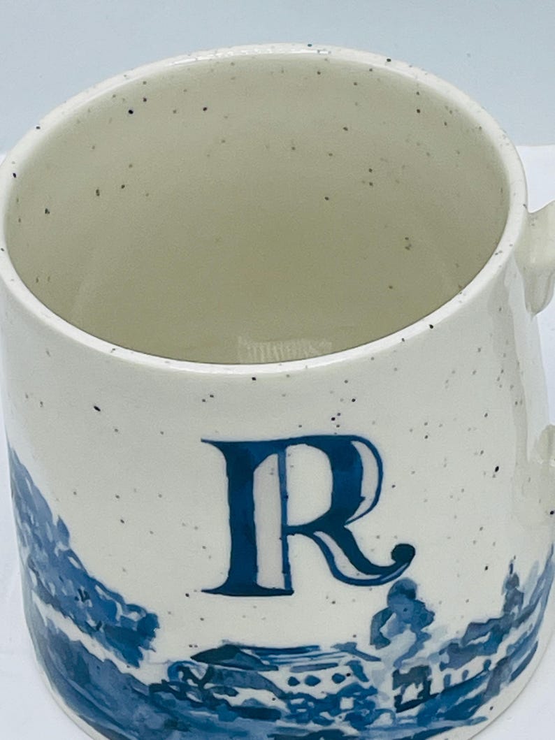 May include: White ceramic mug with a blue letter "R" and a blue landscape design. The mug has a speckled finish and a handle. The design is reminiscent of classic blue and white pottery.
