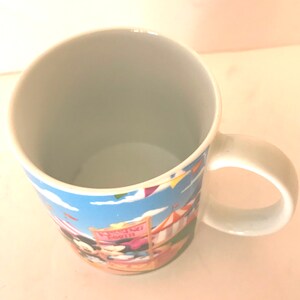 Puede incluir: Una taza de cer&aacute;mica blanca con un dise&ntilde;o de dibujos animados colorido con Mickey Mouse y Minnie Mouse en un carnaval. La taza tiene un fondo de cielo azul con nubes blancas y banderas de colores. El texto "Wishing Well" es visible en un cartel rosa.