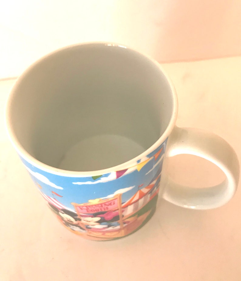 Puede incluir: Una taza de cer&aacute;mica blanca con un dise&ntilde;o de dibujos animados colorido con Mickey Mouse y Minnie Mouse en un carnaval. La taza tiene un fondo de cielo azul con nubes blancas y banderas de colores. El texto "Wishing Well" es visible en un cartel rosa.