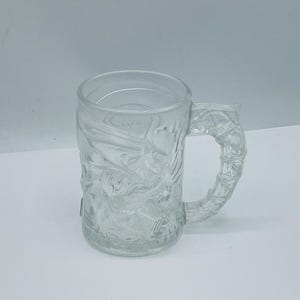 May include: Clear glass mug with a textured design and a handle. The mug has a cylindrical shape with a detailed relief pattern on the exterior. The handle is also textured, and the mug appears to be empty.