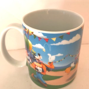 Puede incluir: Una taza de cer&aacute;mica blanca con una imagen de dibujos animados de Goofy sosteniendo un cubo de palomitas de ma&iacute;z. El fondo presenta un cielo azul con nubes blancas y banderas de colores.