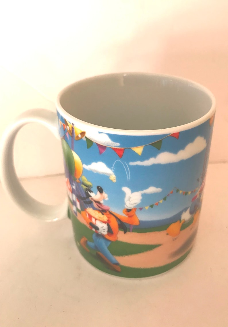 Puede incluir: Una taza de cer&aacute;mica blanca con una imagen de dibujos animados de Goofy sosteniendo un cubo de palomitas de ma&iacute;z. El fondo presenta un cielo azul con nubes blancas y banderas de colores.