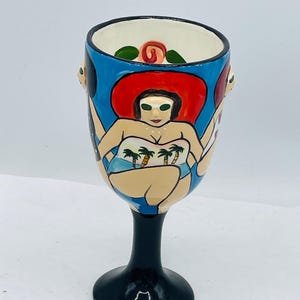 May include: A hand-painted ceramic wine glass featuring a woman in a swimsuit and a red hat. The glass has a black stem and base, with a blue background and a white interior. The design includes a rose and other figures.