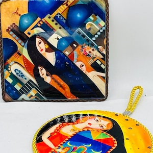 May include: Two decorative ceramic pieces with colorful abstract art. One is square with a brown rope border, featuring figures and architectural elements in blue, orange, and white. The other is round with a yellow rope border and a woman in a colorful dress.