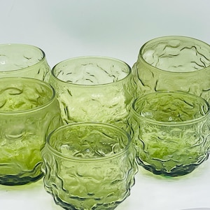 May include: Six green glass tumblers with a textured, organic design. The glasses are a light green color and have a rounded shape. The textured surface gives them a unique, vintage look. Perfect for serving beverages.