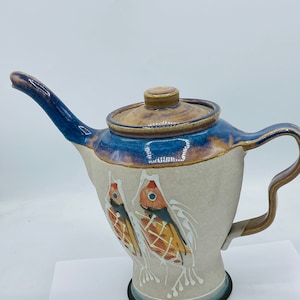 May include: A ceramic teapot with a brown and blue glaze. The teapot has a unique design with two fish painted on the side. The teapot has a lid and a spout.