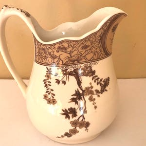 May include: A white ceramic pitcher with a brown floral design. The pitcher has a scalloped rim and a handle.
