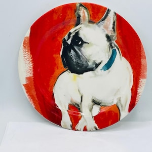 May include: Round ceramic plate featuring a white French Bulldog with a black muzzle and blue collar, set against a vibrant red background. The plate has a glossy finish and is suitable for serving food.