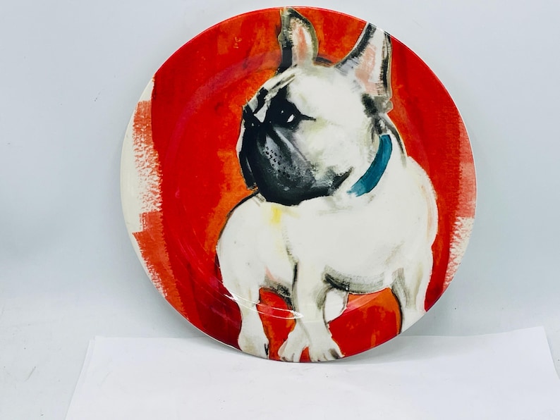 May include: Round ceramic plate featuring a white French Bulldog with a black muzzle and blue collar, set against a vibrant red background. The plate has a glossy finish and is suitable for serving food.