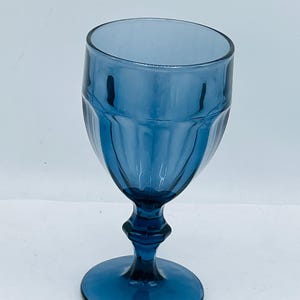May include: A blue-tinted glass goblet with a stem and a round base. The glass has a ribbed design on the bowl and a detailed stem. The goblet is likely used for serving beverages.