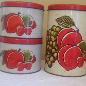 May include: Two vintage metal canisters with red lids and fruit designs. The canisters have a white background with red peaches, green leaves, and red strawberries. The larger canister has a design of grapes and peaches.
