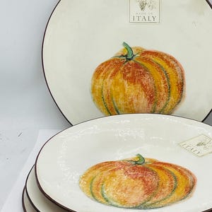May include: A set of cream-colored ceramic plates and a bowl featuring a hand-painted orange and yellow pumpkin design. Each piece has a dark brown rim and a rectangular label that reads Made in Italy with a small grape cluster illustration.