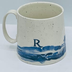 May include: Cream-colored ceramic mug with a blue landscape design and the letter "R". The mug has a speckled finish and a curved handle. The blue design appears to be a stylized depiction of water and a shoreline.
