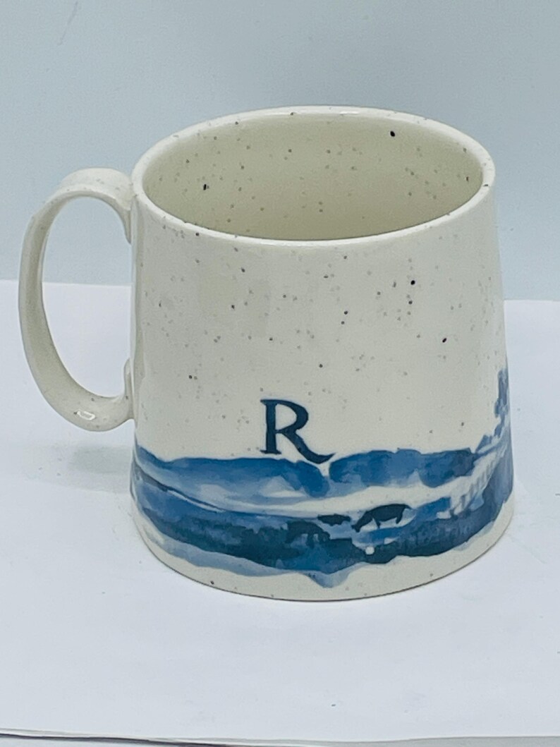May include: Cream-colored ceramic mug with a blue landscape design and the letter "R". The mug has a speckled finish and a curved handle. The blue design appears to be a stylized depiction of water and a shoreline.