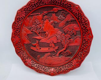 May include: A red lacquered Chinese plate with a carved design of a landscape with mountains, trees, and figures in a boat. The plate has a scalloped edge and a floral border.