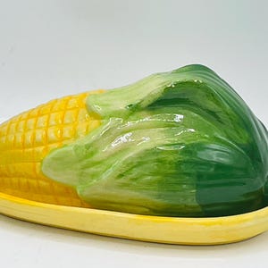 May include: A ceramic butter dish shaped like a corn cob, featuring a yellow base and a lid designed as a yellow corn cob with green husks. The glossy finish highlights the textured kernels and the smooth, curved leaves of the corn cob lid.