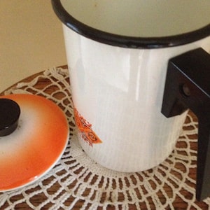 May include: White enamelware coffee pot with a black handle and rim. The pot has an orange and brown design on the side. The lid is orange with a black knob. The pot is sitting on a wooden surface.