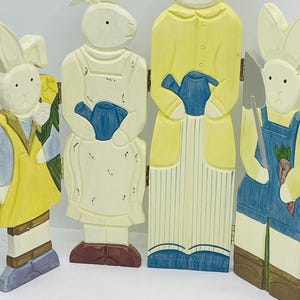 May include: Wooden Easter bunny figurines in yellow, blue, and white. The bunnies are holding a watering can, a shovel, and flowers. The figurines are connected by hinges, creating a decorative Easter display.