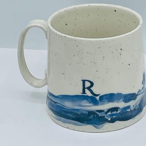 May include: Cream-colored ceramic mug with a blue landscape design and the letter "R". The mug has a speckled finish and a curved handle. The blue design depicts a stylized landscape.