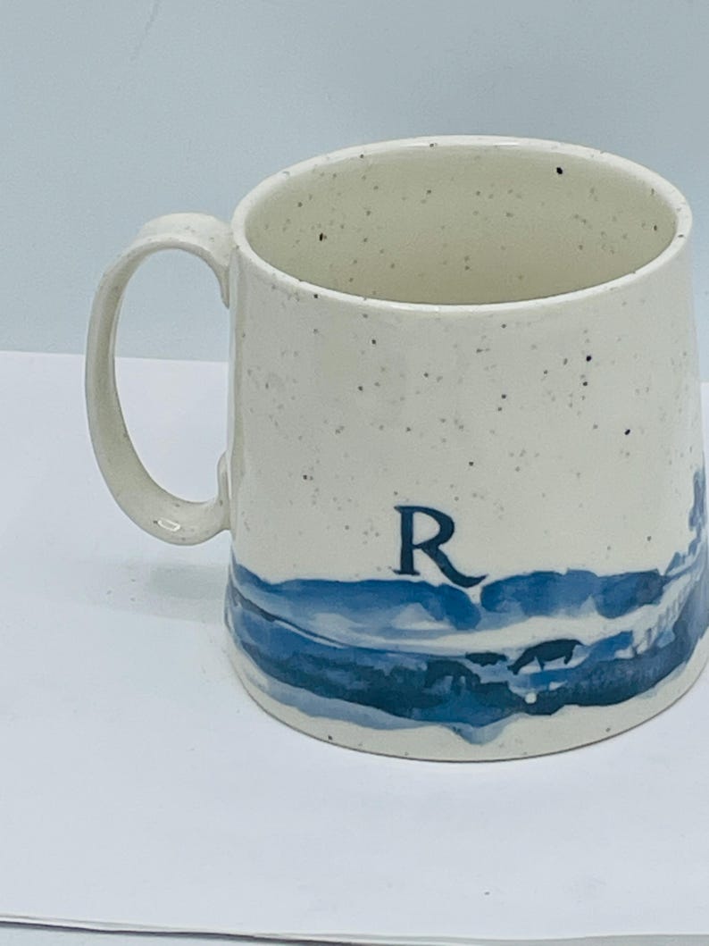 May include: Cream-colored ceramic mug with a blue landscape design and the letter "R". The mug has a speckled finish and a curved handle. The blue design depicts a stylized landscape.