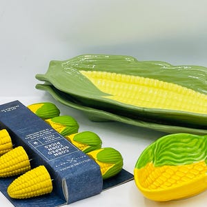May include: A set of ceramic corn-shaped serving dishes and corn-shaped picks. The dishes are yellow with green husks. The picks are yellow corn kernels with green husks, packaged in a blue box labeled Corn Shaped Corn Picks.